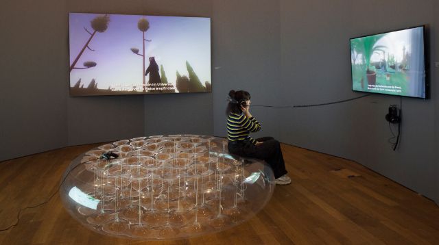 Ausstellungsansicht game design today, eine frau sitzt auf einem transparenten gefäss, mit aufgesetzten kopfhörern 