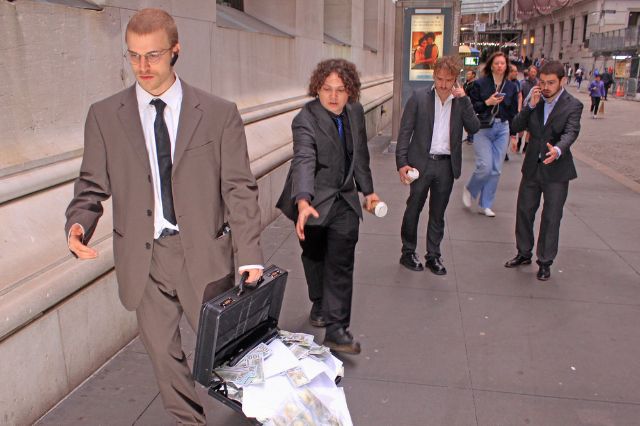 4 junge Männer in Anzügen und einem offenen Koffer voll Geld laufen einer Strasse in Manhattan entlang