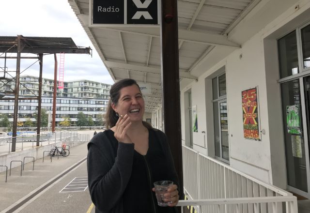 Eine Frau lacht, hält ein Wasserglas und eine Zigarette in den Händen. Im Hintergrund ist links der Eingang von Radio X.