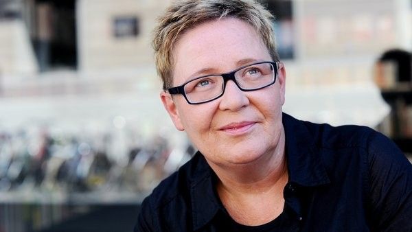 Portrait von einem weiblich gelesenen Gesicht mit eckiger Brille, blonden, kurzen Haaren und einem Lächeln.