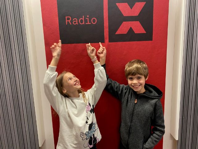 Zwei Kinder im Studio von Radio X zeigen auf dessen Logo 