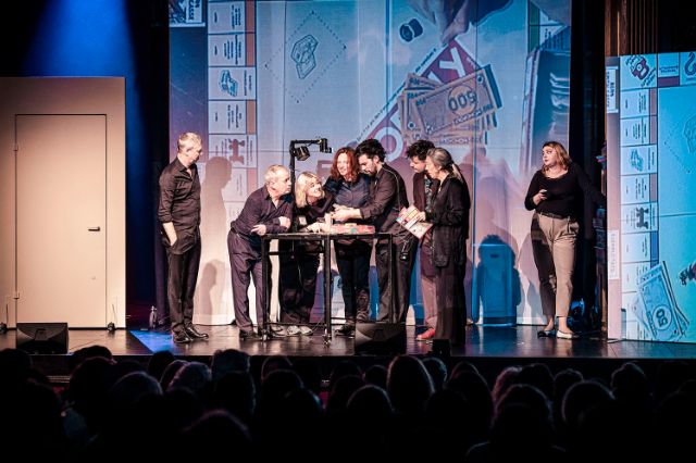 Acht Menschen auf einer Bühne, ein Monopoly-Spiel wird projiziert, Publikum im dunklen Vordergrund