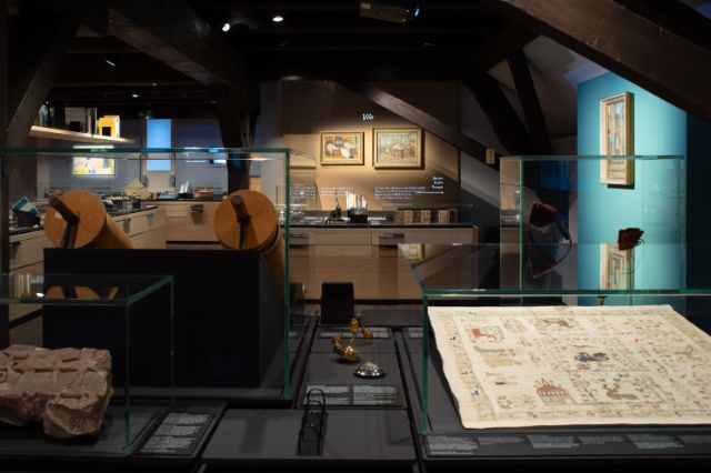 Ein Ausstellungsraum mit Vitrinen, Bilderrahmen auf einer türkis Wand, dunkle Balken