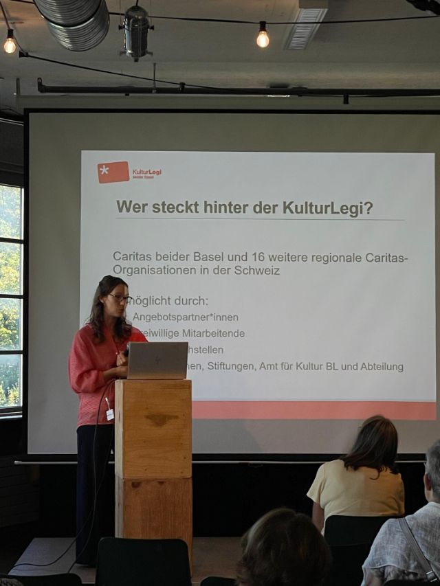 Ein Referat mit einer projizierten Folie im Hintergrund, Zuhörende von hinten fotografiert.