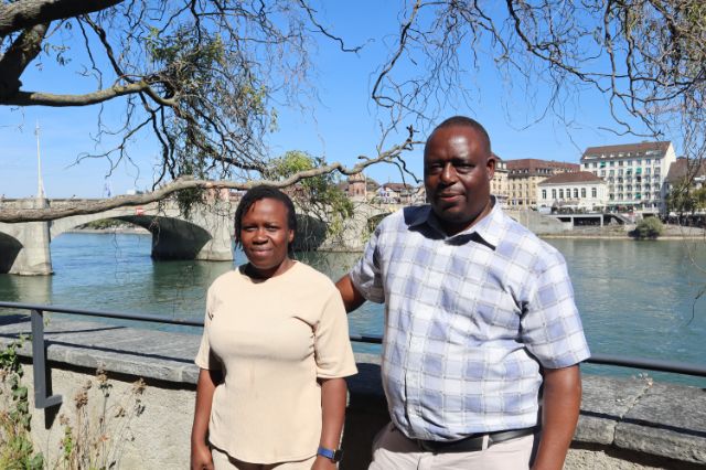 A woman and a man from Zimbabwe stand next to each other in the foreground, the background is the mittlere Brücke in Basel.