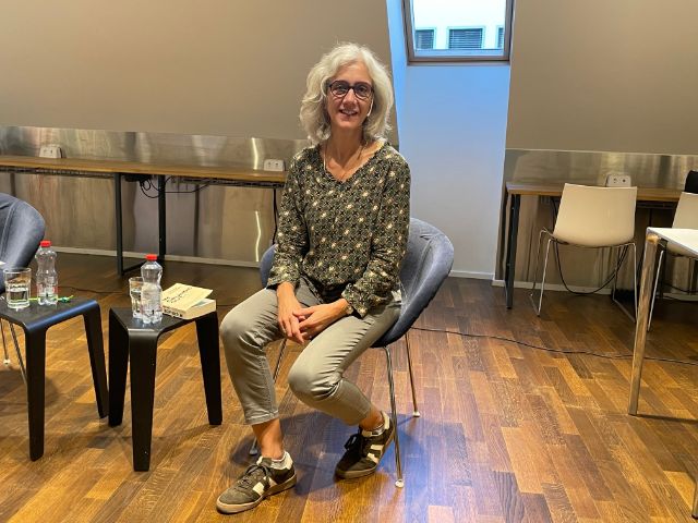 Eine Frau mit dunkler Brille, halblangen Haaren und gemusterter Brille sitzt in einem Sessel