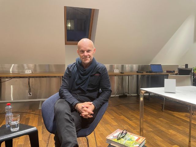 Ein schwarz gekleideter Mann sitzt lächelnd auf einem Sessel, im Vordergrund ein Buch und Lesebrille