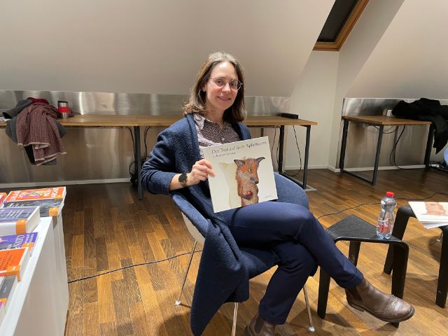 Eine Frau mit Brille und einem Buch vor dem Oberkörper lächelt in die Kamera