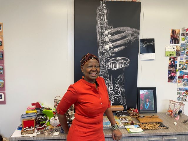 Eine Frau in rotem Kleid steht vor einem Tisch mit Schmuck und einem Bild mit einem Saxophon