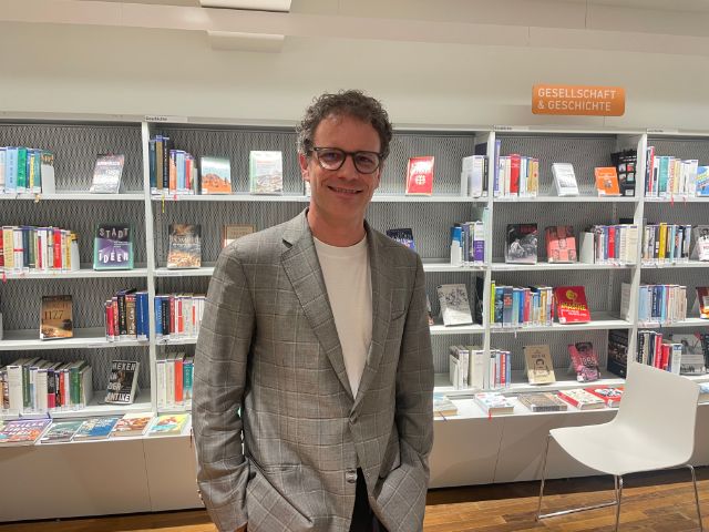 Ein Mann mit Brille und Kittel lächelt vor einem Buchregal in der Bibliothek 