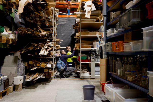 Viele Regale voll mit Brettern, Boxen, Eimer und Material. Im Hintergrund ein erwachsener Mensch und ein Kind