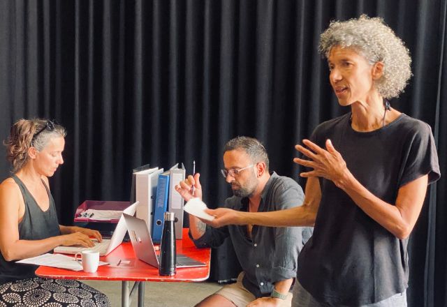 Drei Personen auf einer Bühne: links sitzen zwei Menschen an einem Schreibtisch, rechts spricht und gestikuliert eine Person mit Geprächsnotizen in der Hand