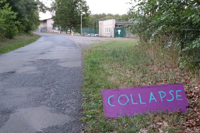 Geteerte Strasse mit einer Wiese auf dem ein Schild steht, auf dem Collaps steht. 