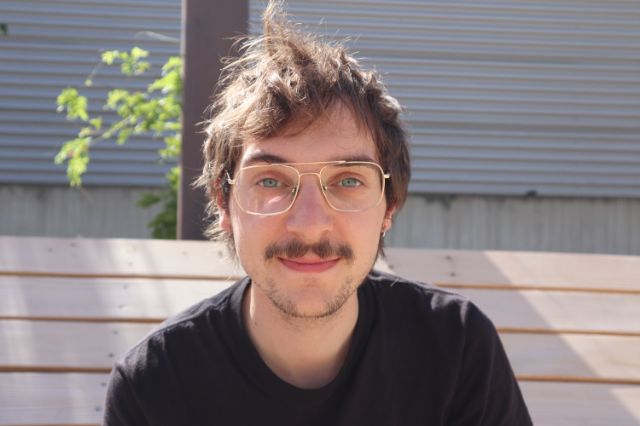 Ein Porträt von Laurel Bloom. Er hat hellbraunes Haar, blaue Augen und einen kleinen Schnauzbart. Er trägt eine grosse, dünne Brille und ein schwarzes T-Shirt. Er lächelt scheu in die Kamera.