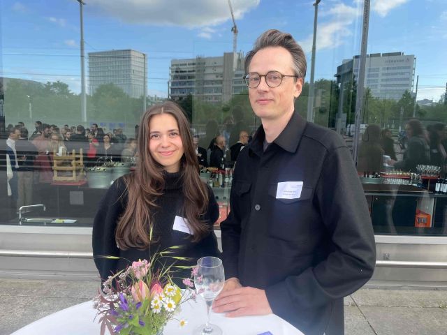 zwei schwarz bekleidete Menschen stehen an einer Spiegelfassade, im Vordergrund ein Blumenstrauss