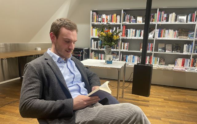 Ein Mann sitzt mit einem Buch in der Hand in der Bibliothek, Beine überschlagen