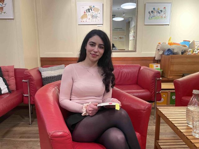 Eine junge Frau mit langen Haaren sitzt in einem Sessel, hat ein Buch im Schoss und lächelt