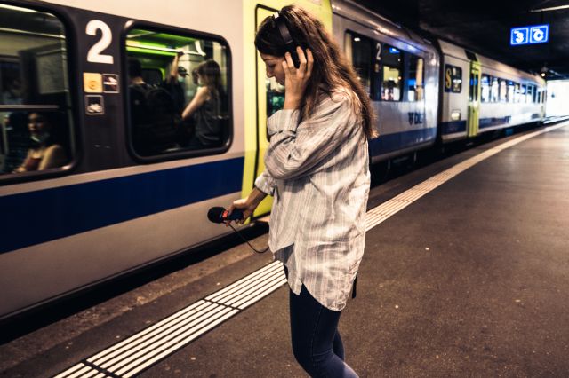 Eine Frau mit Mikrofon und Kopfhörern steht auf einem Zugperron. Vor ihr ein voller Zug.