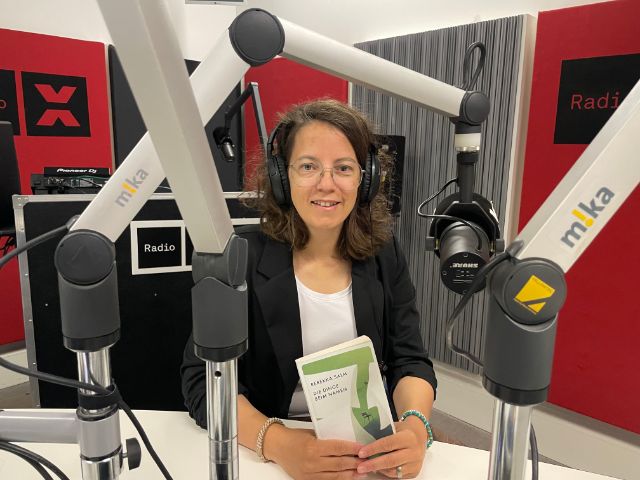 Eine Frau mit Brille und dunklen halblangen Haaren sitzt im Studio, sie hält ein Buch vor sich