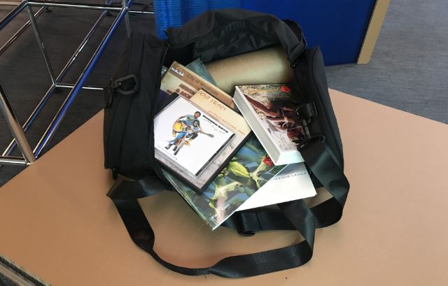 Eine geöffnete Reisetasche zeigt Gegenstände wie DVD, CDs, Broschüren und Plakatrolle