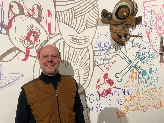 Ein Mann lächelt in die Kamera vor einem Wandgemälde mit runden Charaktergesichtern und einer Maske