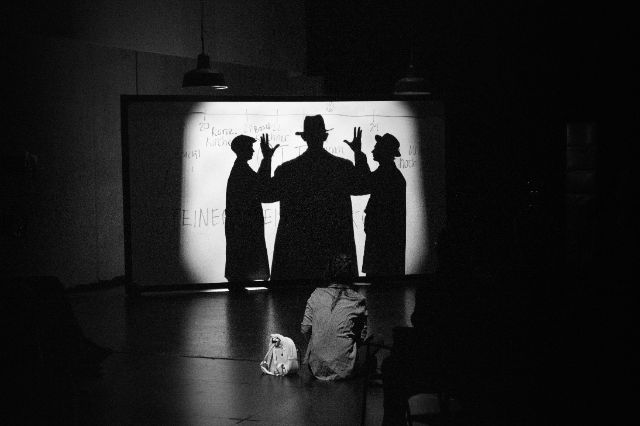 Schattenspiel an einer beleuchteten Wand, drei Figuren mit Hüten und erhobenen Händen