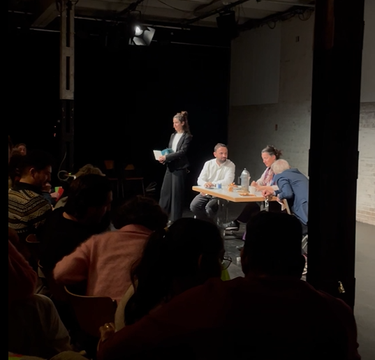 eine beleuchtete Theaterbühne mit drei Menschen an einem Tisch und einer stehenden Sprecherin