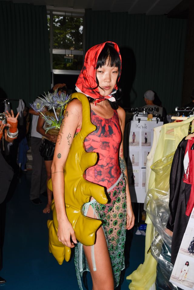 Ein Model steht im Backstage und trägt eine gelbe Tasche und ein rotes Kopftuch. 