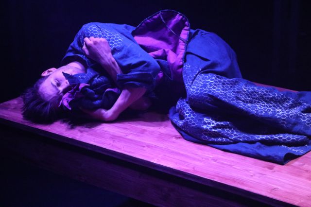 Szene aus dem stück Stimmenmeer, ein mann in blau gekleidet liegt am Boden, in den armen ein Tuch.