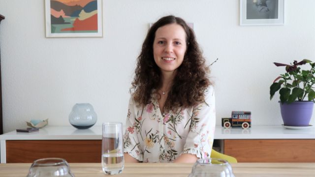 Eugenia Senik sitz an einem Tisch in einem Haus. Im Hintergrund sind Vasen, Bilder und Pflanzen.