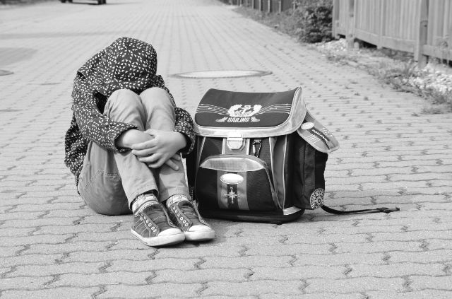 Ein Kind mit hochgezogener Kapuze und Schulsack sitzt am Boden