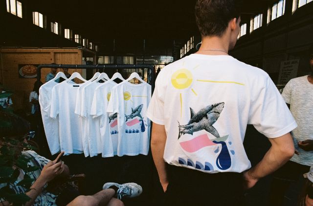 Mann mit auffälligem T-Shirt mit Sonne und Hai, im Hintergrund gleiches T-Shirt auf Kleiderbügel