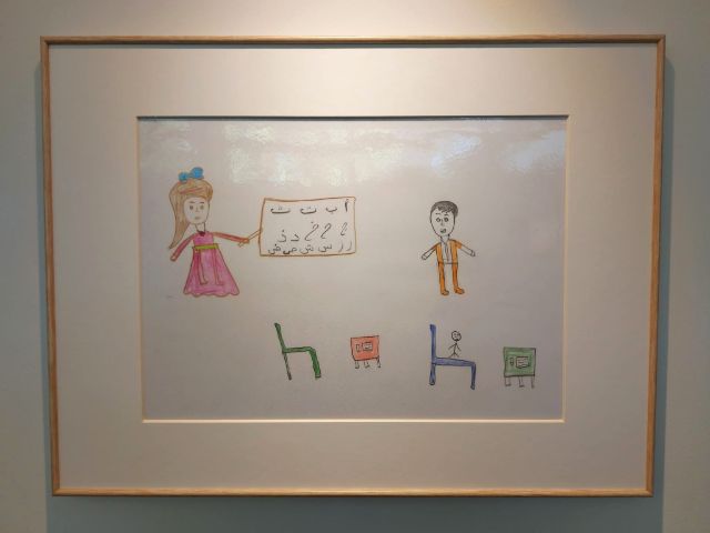 eine der elf ausgestetllten kinderzeichnungen. wir sehen eine lehrerin, stuehle, ein kind und arabische schriftzeichen. 