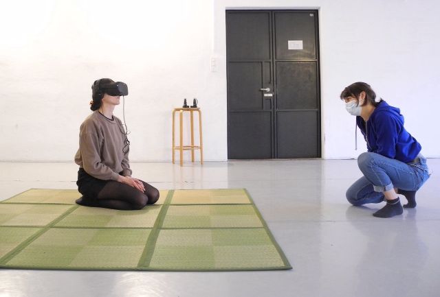 Person mit einer VR-Brille ist auf ihren Knien und wird von einer anderen Person beobachtet.
