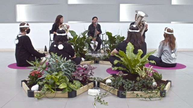Im Hintergrund spielen drei Musiker:innen auf Blasinstrumenten; Vor den Musiker:innen sitzen vier Personen, sie bilden mit den Musiker:innen einen Kreis und haben uns den Rücken zugewandt, sie tragen ein Messgerät auf dem Kopf; Töpfe mit Pflanzen stehen im Raum verteilt