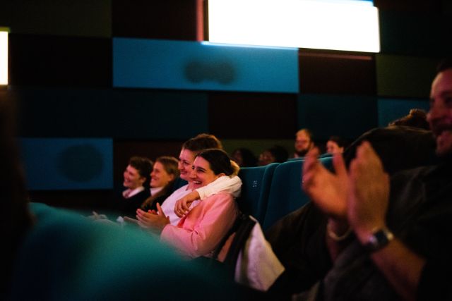 Man sieht applaudierende Menschen in einem Kino, im Fokus sind zwei Frauen, die aneinanderlehnen.