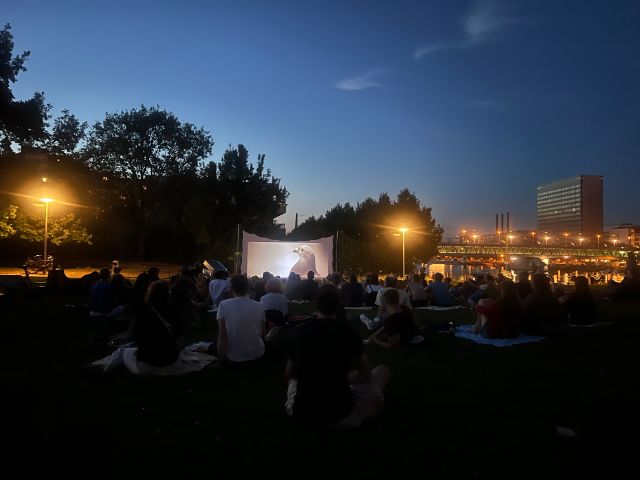 Bild zeigt Filmpublikum, welches auf Decken im St.Johanns-Park sitzt. Vorne wird ein Film auf eien Leinwand projeziert.