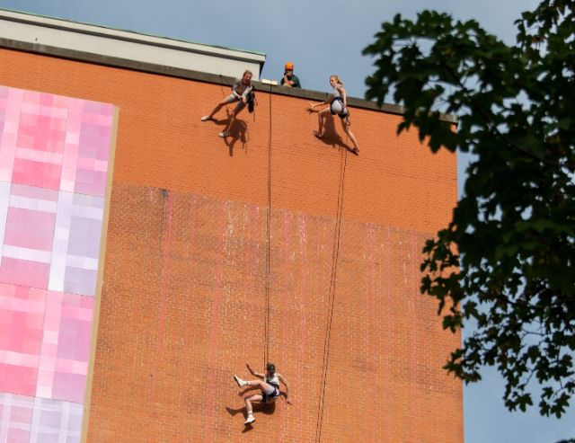 drei Artist:innen hängen abgeseilt an einer grossen Backstein-Fassade