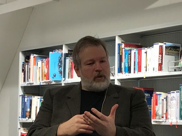 Ein sprechender Mann gestikuliert, im Hintergrund Regale der Bibliothek
