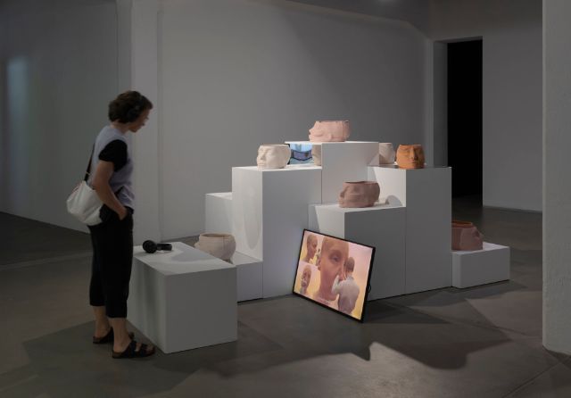 Eine Frau blickt in einem Ausstellungsraum auf einem Bildschirm. Dort herum ausgestellt sind Keramiken von Gesichtern.