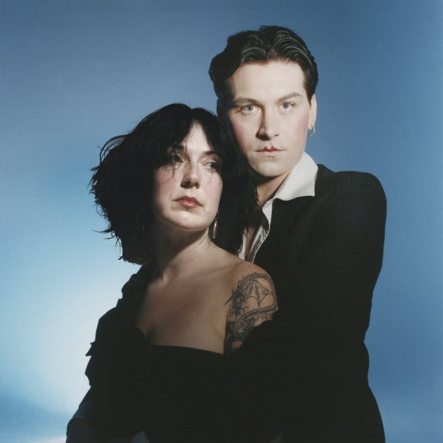 Ein bleiches, in schwarz-weiss gekleidetes Paar posiert vor blauem Hintergrund