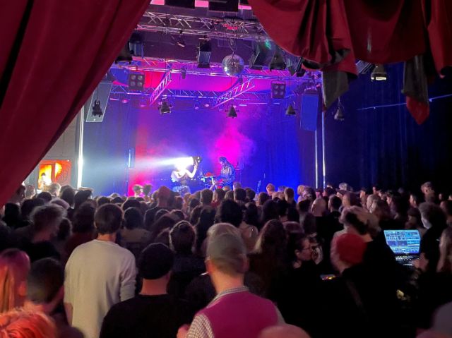 das bild zeigt einen gefüllten konzertsaal mit 2 performenden Personen auf der Bühne