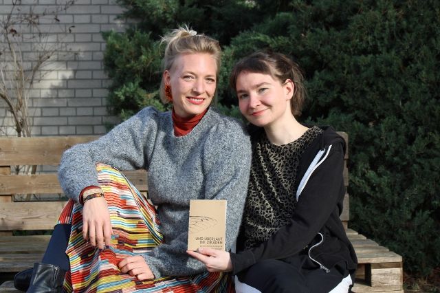 Valerie Meyer und Julie Rüegger sitzen lächelnd auf einer Holzpallette im Freien. Sie halten ihr Buch: "Und überlaut die Zikaden"