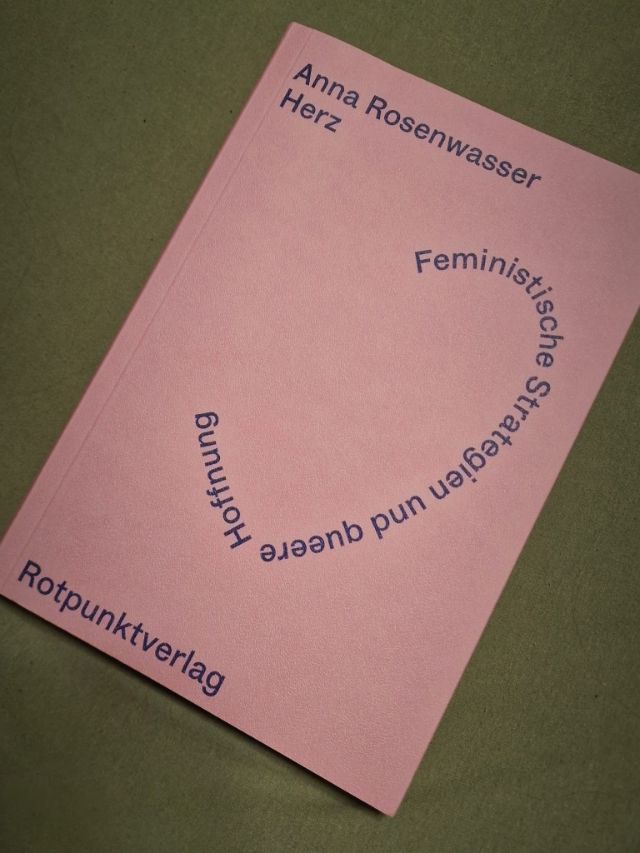 Das Buch auf grünem Hintergrund. Das Buch ist rosa, darauf ist in blauer Schrift der Titel in Form eines Herzens