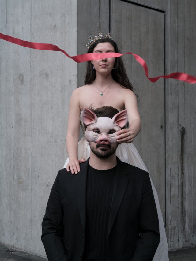 Eine Frau im rosa Prinzessinnenkleid und Krone hält eine Schweinemaske vor dem Gesicht eines schwarzgekleideten Mannes, der vor ihr kniet. Beide schauen gerade nach vorne. Ein rotes Band verdeckt die Augen der Prinzessin.