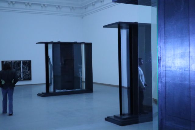 zeigt den gedimmten Ausstellungsraum mit den Skulpturen von Marie Matusz. Sie sind kastenförmig, es spiegeln sich Personen im Plexiglas, das Material ist dunkel.