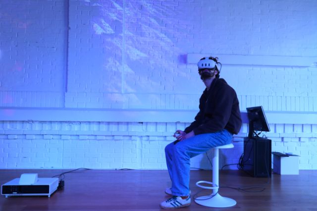 Eine männlich gelesene Person mit VR Brille sitzt in einem Ausstellungsraum