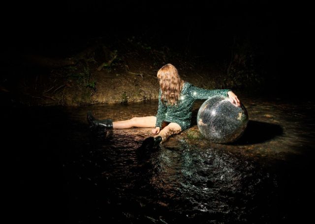 Eine Frau in grünem Glitzerkleid sitzt im Dunkeln in der Natur. Eine Diskokugel ist neben ihr, sie stützt ihren linken Arm darauf ab. Das Gesicht ist weggedreht und von langen, braunen Haaren verdeckt.