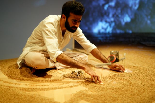 Abhushek Thapar sitzt in weisser Kleidung im Schneidersitz, er bewegt kleine Schilder durch den weizenbedeckten Boden. Im Hintergrund eine Videoprojektion.