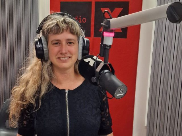 Foto von Sarah Horst im Radio Studio. Sie hat lange blonde gewellte Haare mit einem Pony und eine blaue Bluse. Sie lächelt in die Kamera, während sie Kopfhörer trägt und das Mikrofon vor ihr zu sehen ist. Im Hintergrund ist das Radio X Logo auf einem roten Studiopanel.